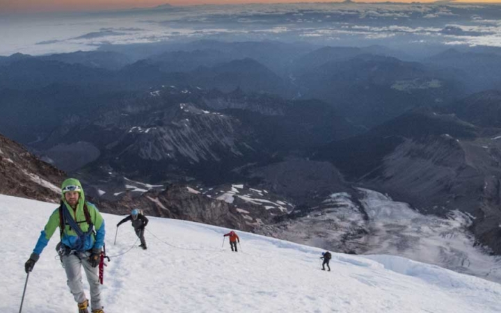 High above a cast mountainous landscape, four people wearing safety gear make their way up a snowy incline.
